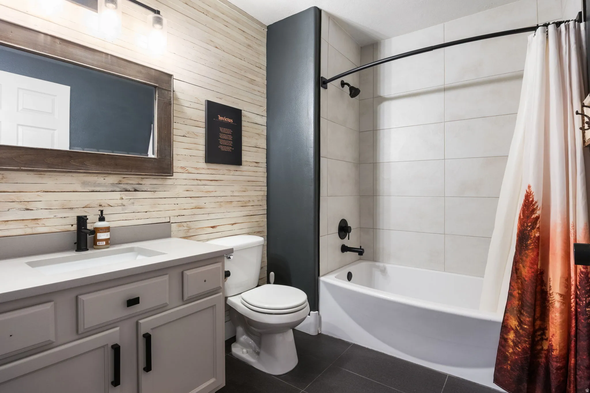 Bathroom featuring a light wood-paneled accent wall, a white vanity with a light countertop and black hardware, a black-framed mirror, and a full-size bathtub with white tile surround and matte black fixtures