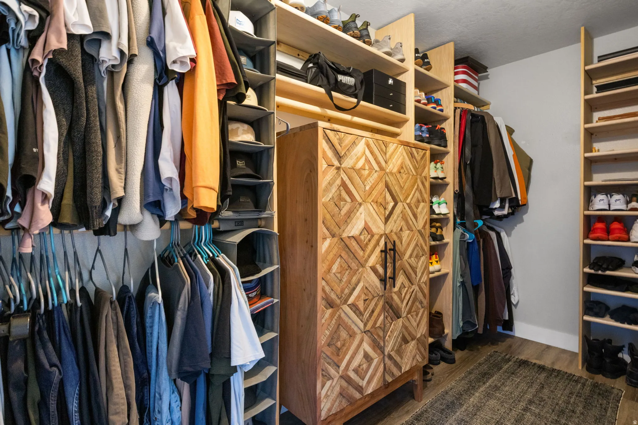 Custom-built closet system featuring extensive shelving, multiple hanging rods, a central wood-finish storage cabinet with geometric door panels, and durable wood-finish flooring