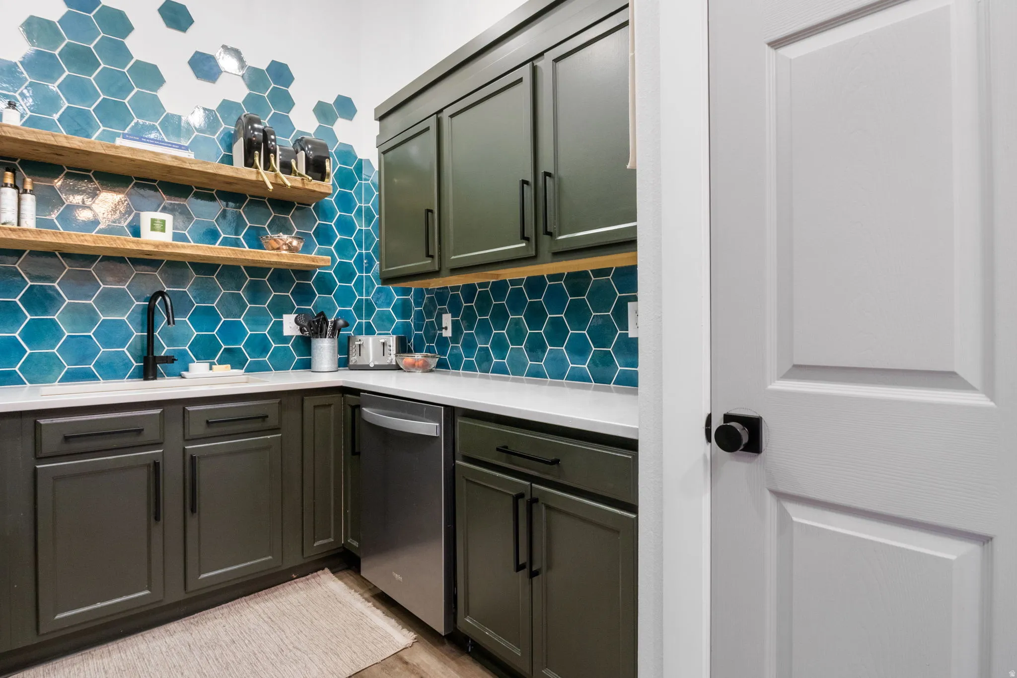 Kitchenette with a vibrant blue hexagonal tile backsplash, dark green cabinetry, white countertops, and a stainless steel dishwasher