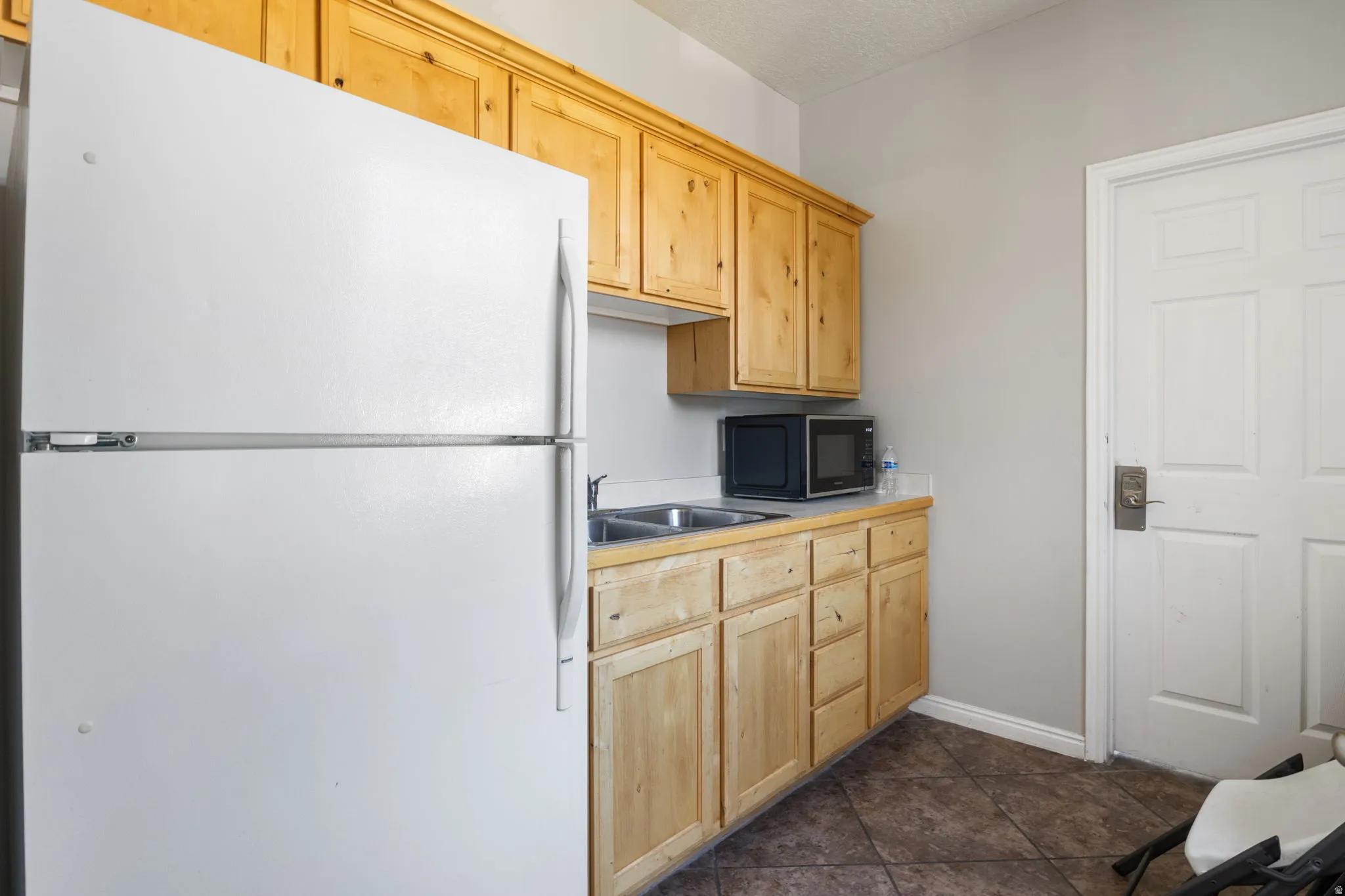Full-size refrigerator, wood-finish cabinetry, dual-basin stainless steel sink, solid surface countertop, and durable tile flooring