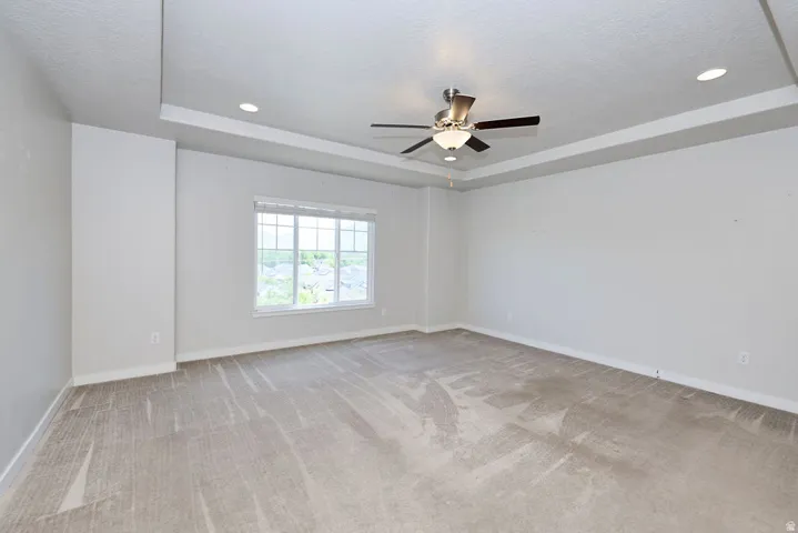 Spacious room featuring neutral carpeting, a large multipane window, a tray ceiling with recessed lighting, and a ceiling fan with integrated lighting