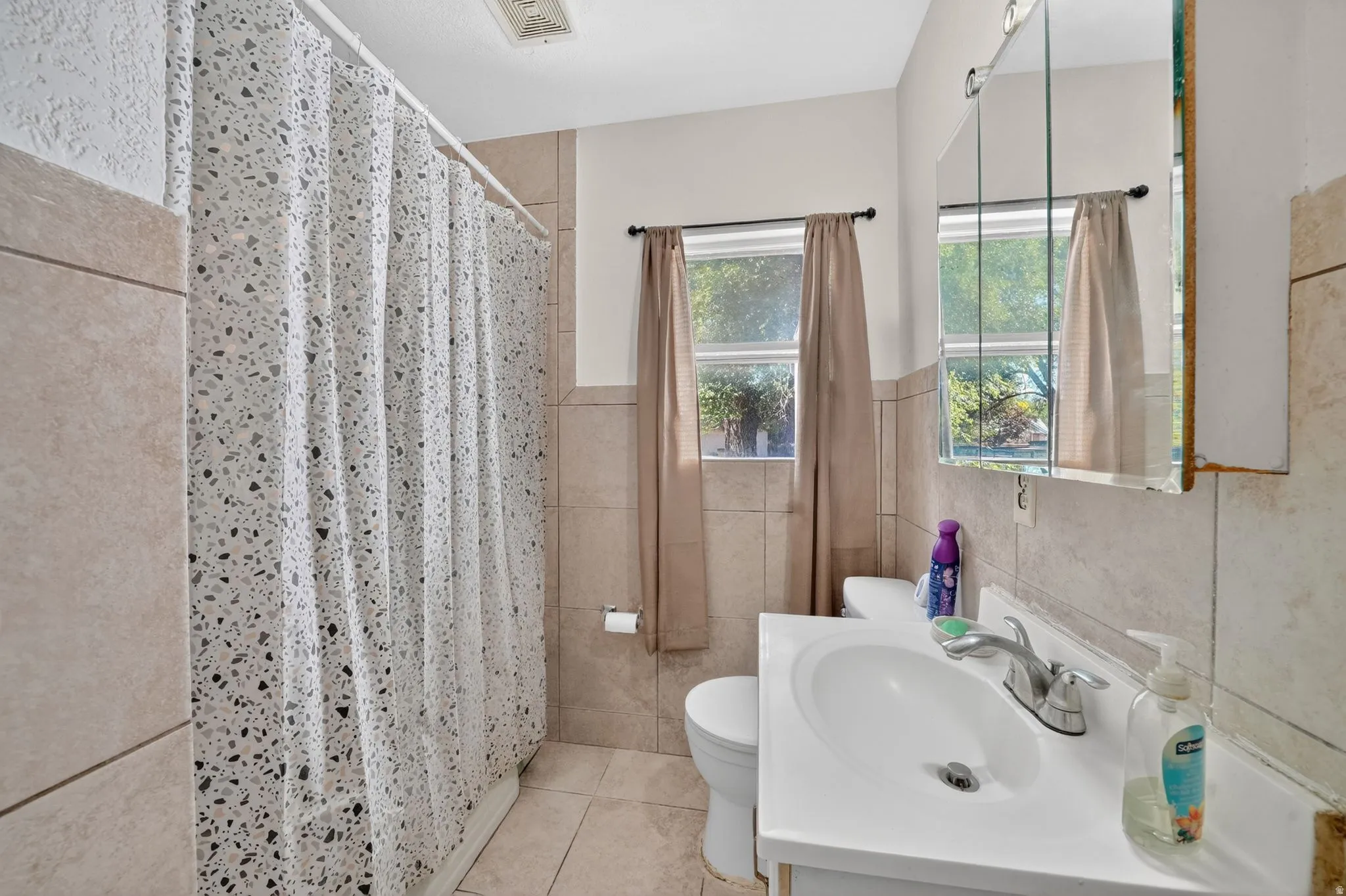 Bathroom featuring tiled walls, a window with natural light, a white vanity with an integrated sink, a mirrored medicine cabinet, and tiled flooring