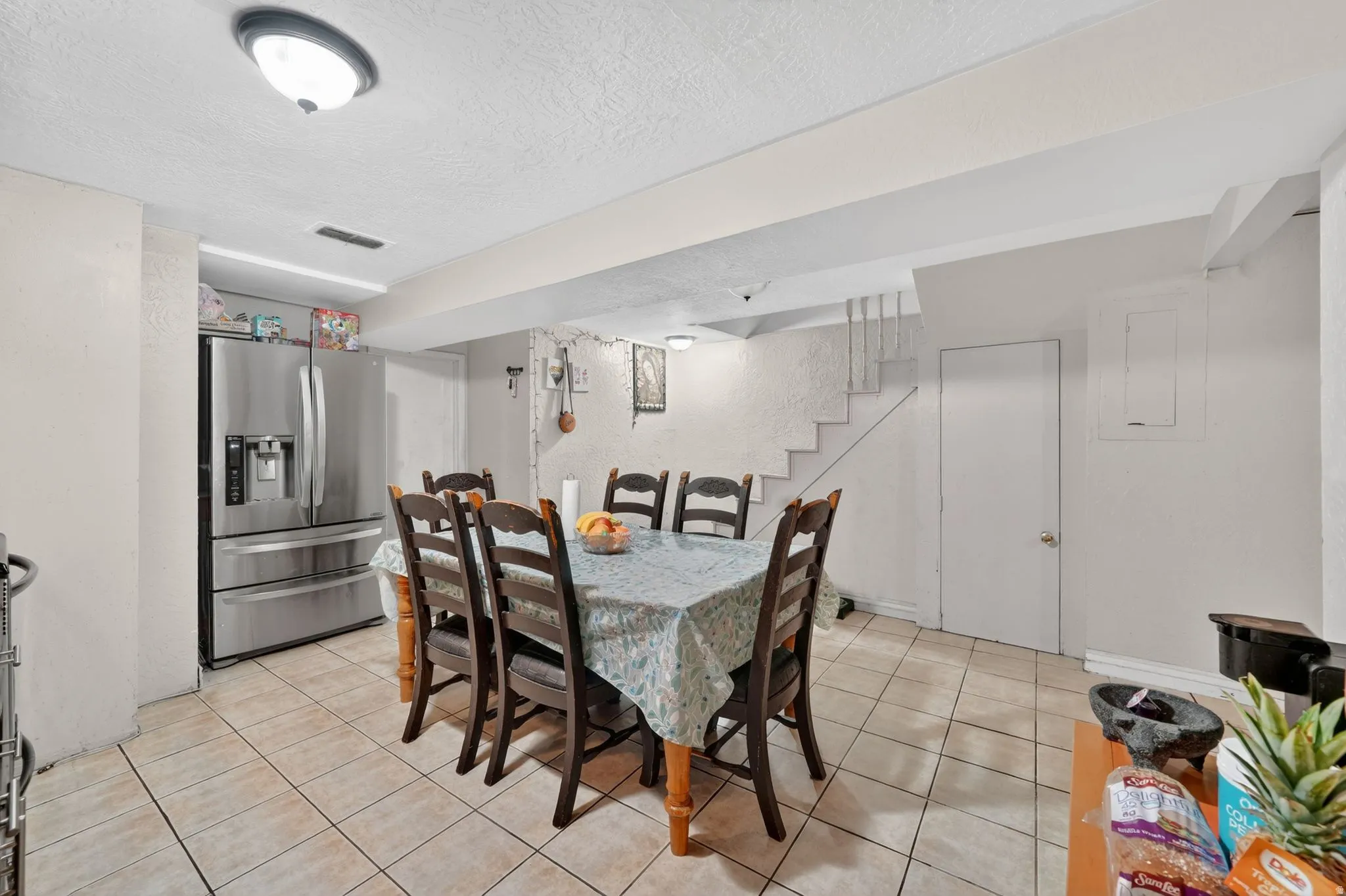 Spacious dining area featuring neutral-toned tile flooring, a built-in stainless steel refrigerator, and a staircase with a white railing