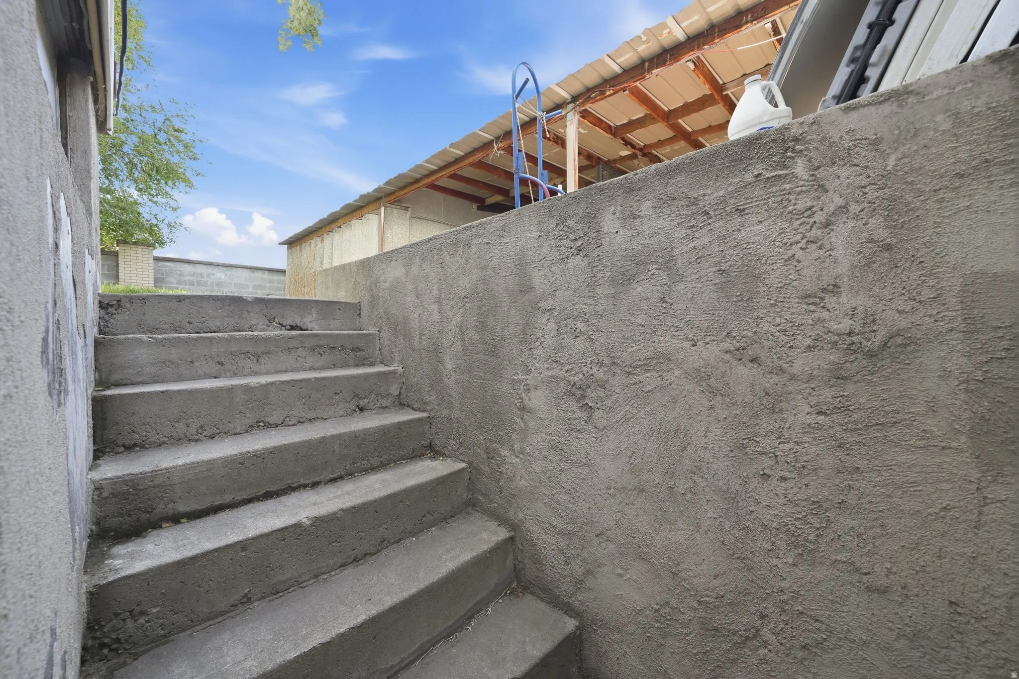 Concrete steps ascending alongside a textured concrete wall, with a partial view of a covered structure and blue sky above