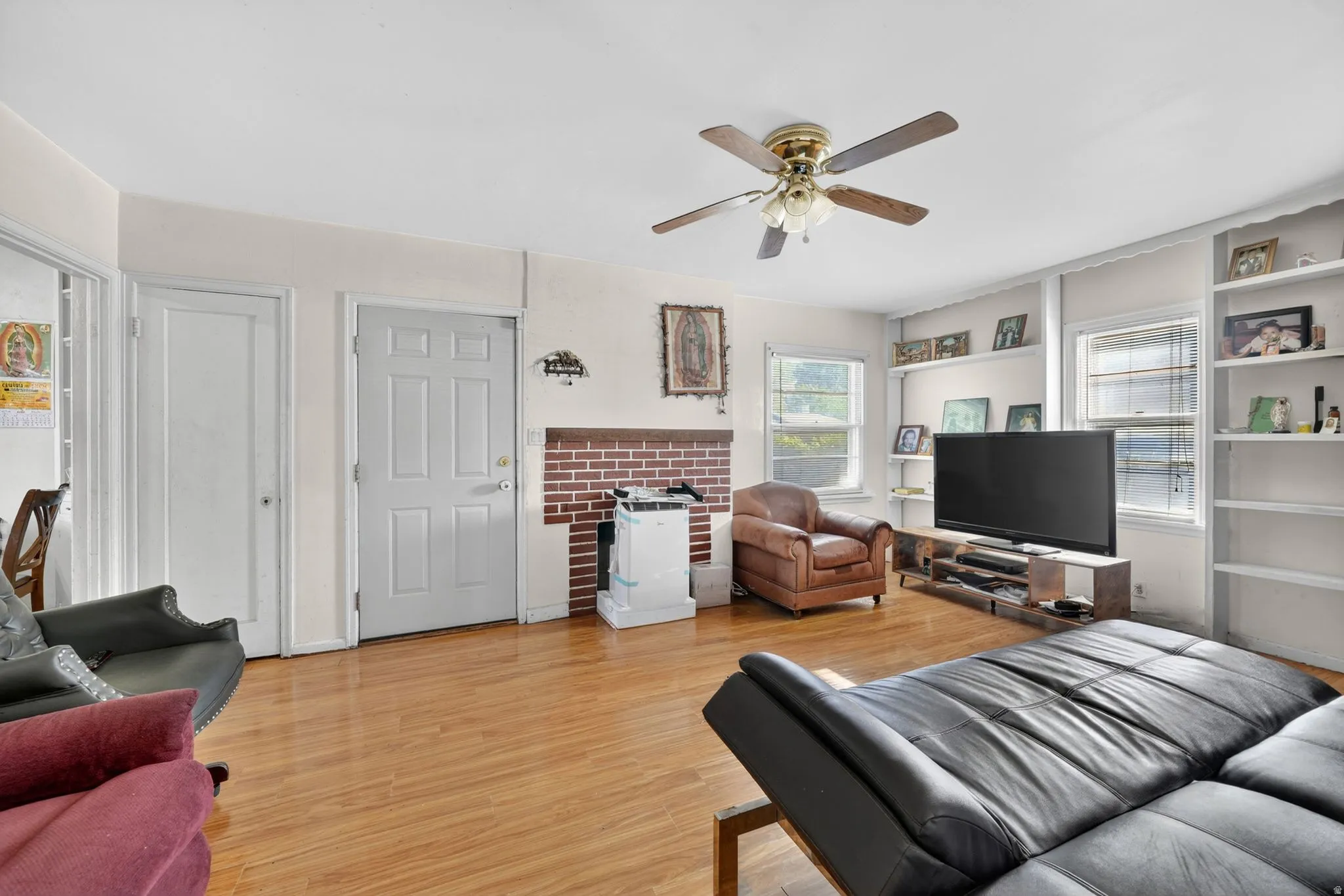 Living area featuring wood-finish flooring, a brick fireplace surround, built-in shelving, multiple windows, and a ceiling fan with integrated lighting