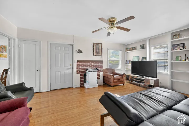 Living area featuring wood-finish flooring, a brick fireplace surround, built-in shelving, multiple windows, and a ceiling fan with integrated lighting