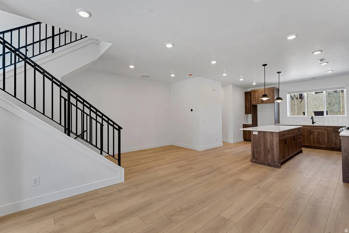 Open-concept living area featuring light wood-finish flooring, a modern kitchen with a large island, sleek dark cabinetry, white subway tile backsplash, and contemporary pendant lighting