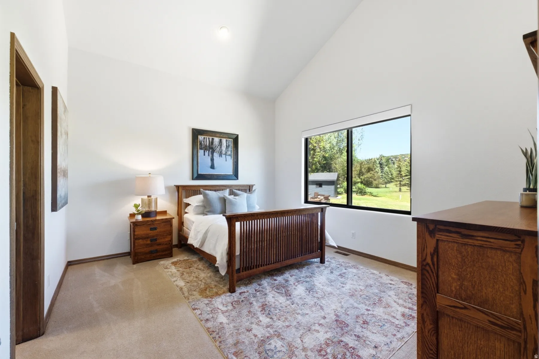 Vaulted ceiling bedroom with a large picture window, light carpeting, and wood-finish trim