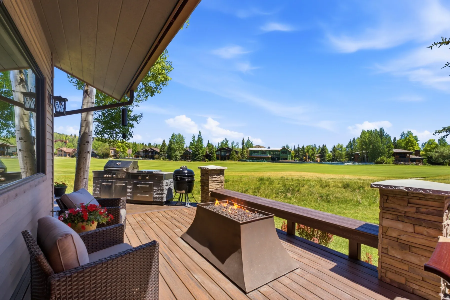 Expansive outdoor deck featuring wood-finish flooring, a built-in outdoor kitchen with stainless steel appliances, a stone-clad fire pit, a built-in wood bench, and stone-clad pillars