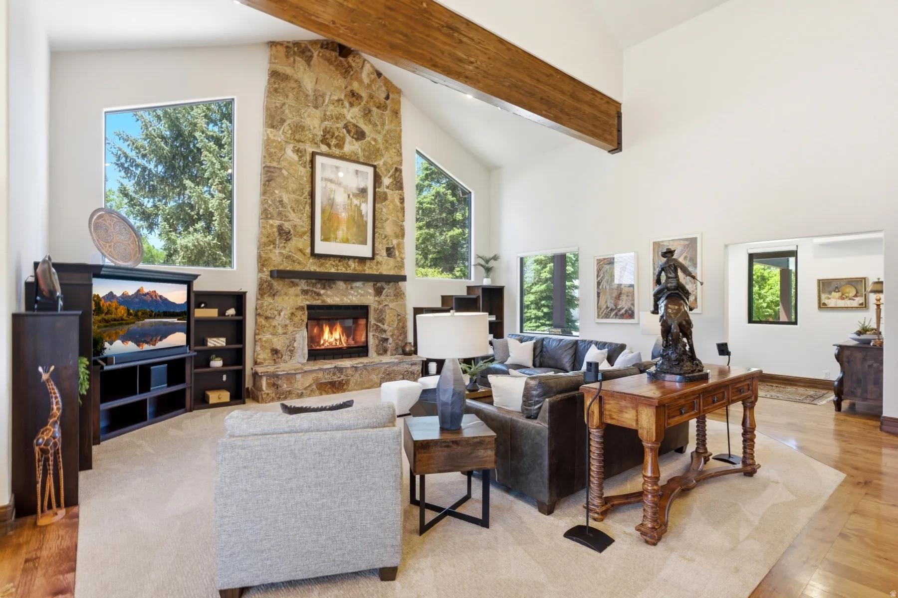 Spacious living area featuring a stone-clad fireplace, vaulted ceilings with exposed wood beams, large windows, wood-finish flooring, and light-toned carpeting