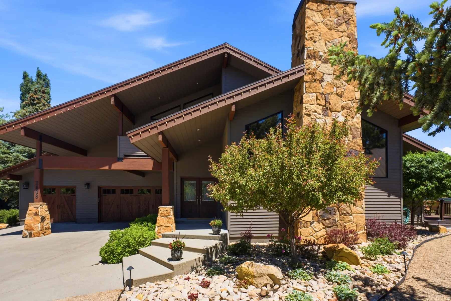 Modern exterior featuring a stone chimney, wood-finish garage doors, concrete steps, and a landscaped rock garden