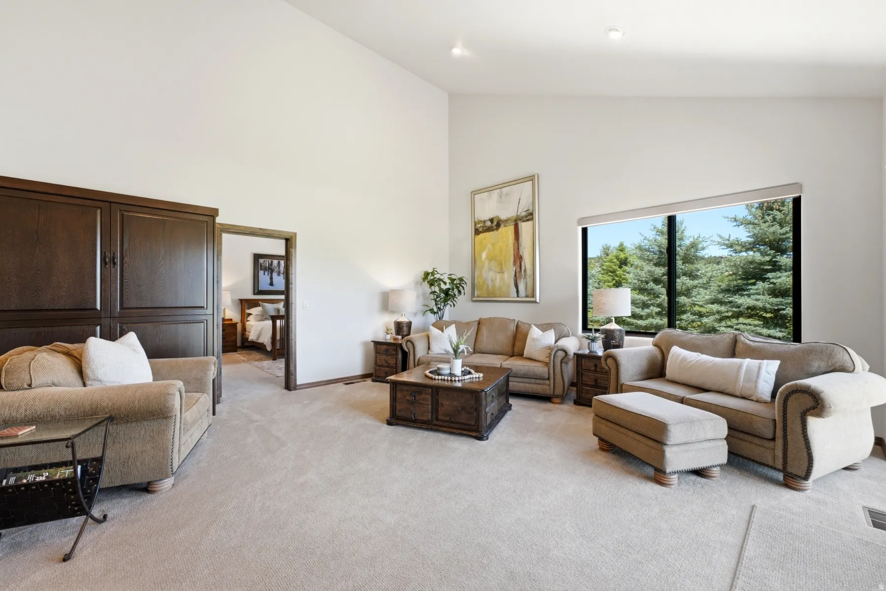 Spacious living area featuring a high vaulted ceiling, large picture window with tree scenery, light-toned carpeting, recessed lighting, and a built-in dark wood storage unit