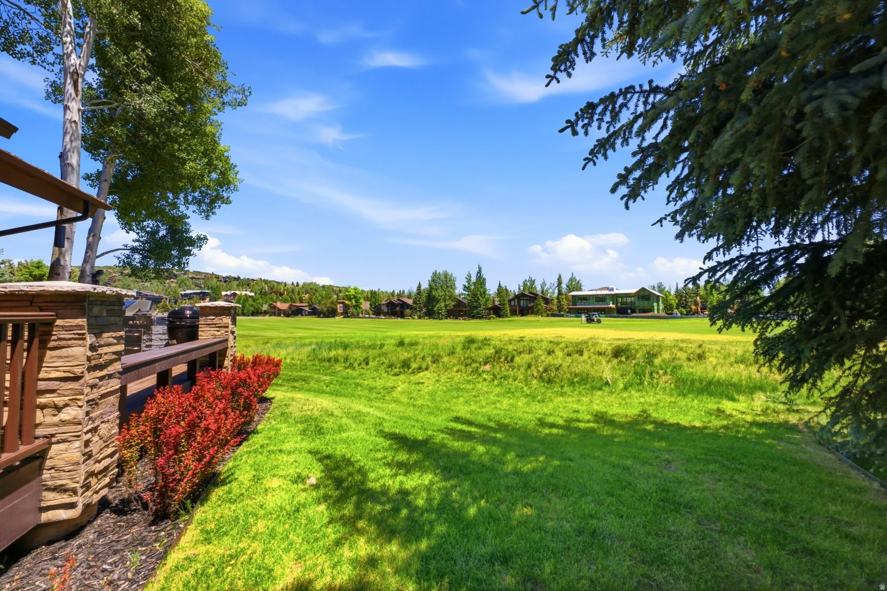 Expansive green space featuring a wood-finish deck with stone-clad pillars, a dark wood-finish railing, and mature landscaping