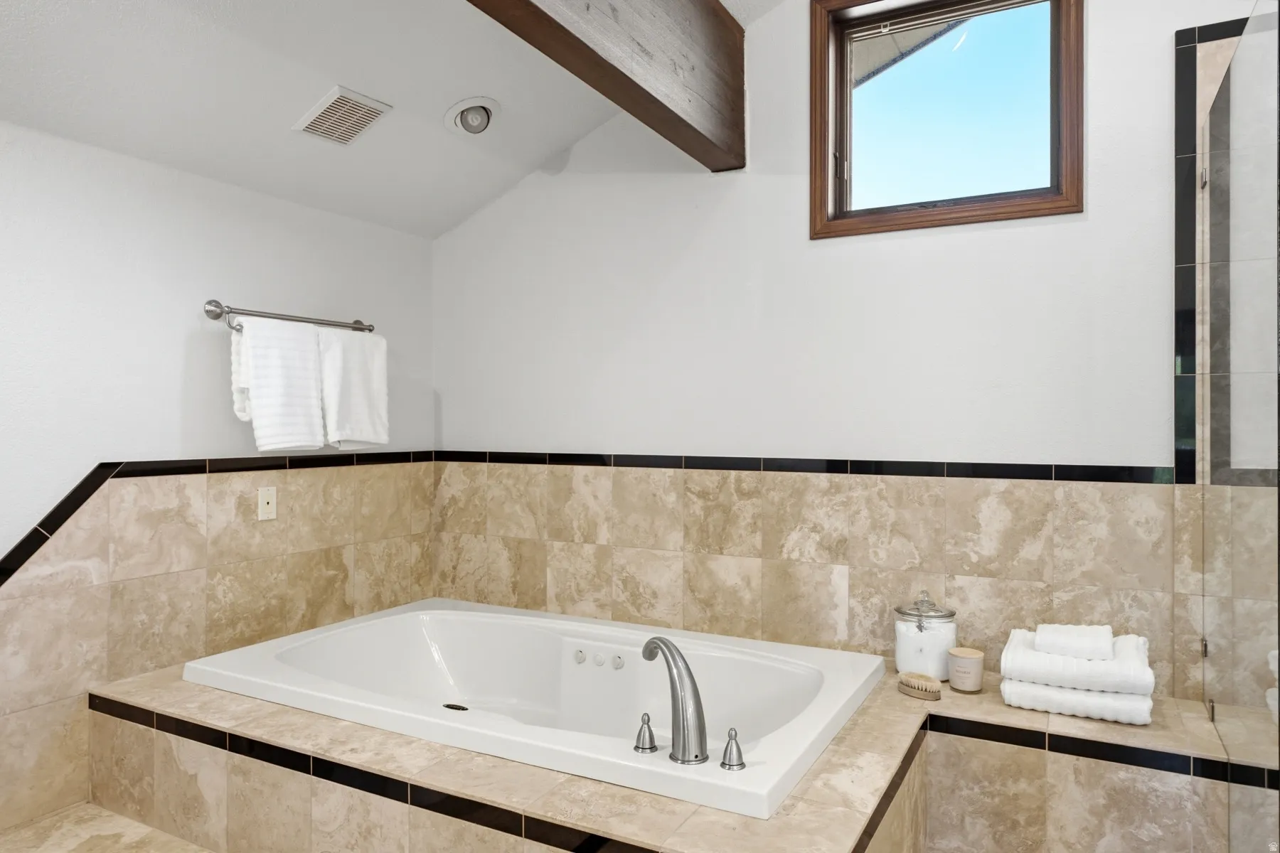 Built-in bathtub featuring a deck-mounted faucet and surround finished with light-toned stone tile and a dark accent stripe