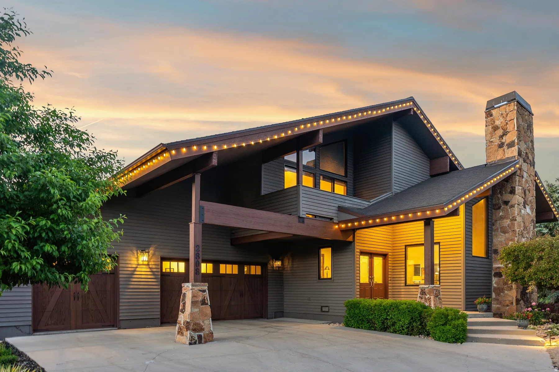 Modern architectural dwelling featuring a stone chimney, extensive window placement, a covered entry, and a two-car garage with wood-finish doors
