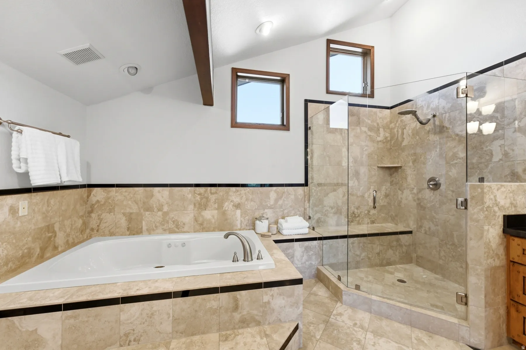 Spacious bathroom featuring a built-in jetted tub, a glass-enclosed shower with a built-in bench, natural stone tile flooring and wall surrounds, exposed wood ceiling beams, and dual high-set windows
