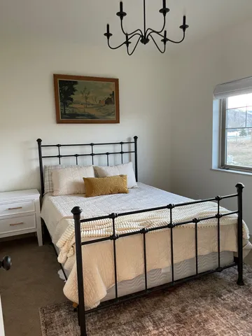 Light-filled room featuring a black wrought iron bed frame, a white two-drawer nightstand with gold-tone hardware, and a black chandelier