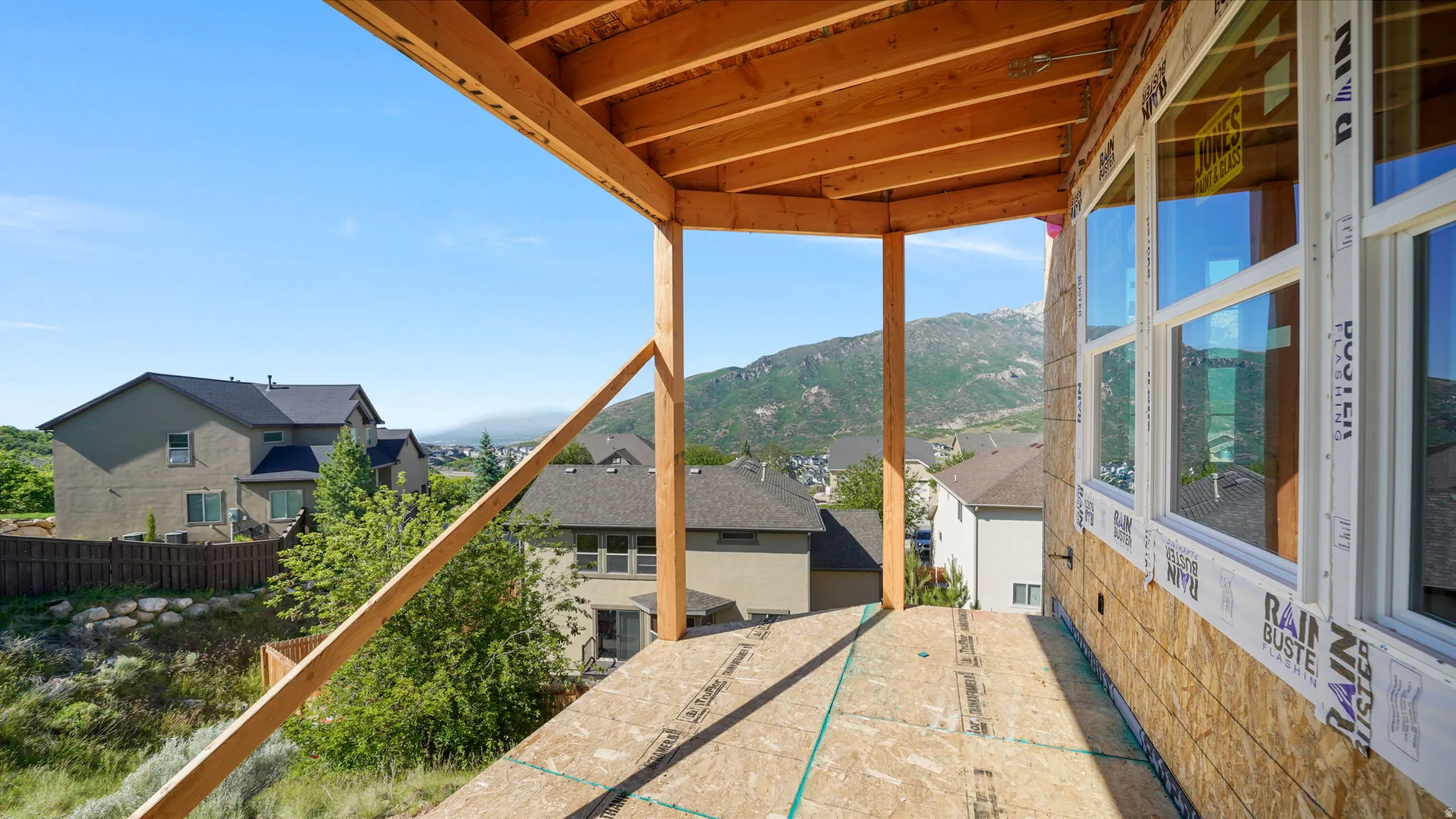 Covered exterior deck with exposed wood framing and sheathing, featuring expansive windows and mountain scenery
