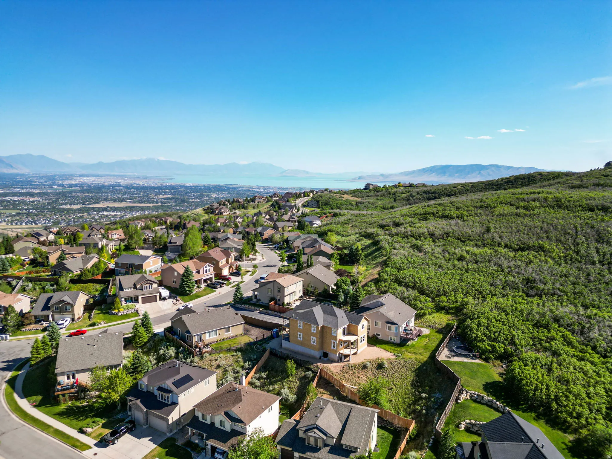 Aerial perspective showcasing a residential neighborhood with varied architectural styles, nestled against a backdrop of distant mountains, a body of water, and extensive green hills