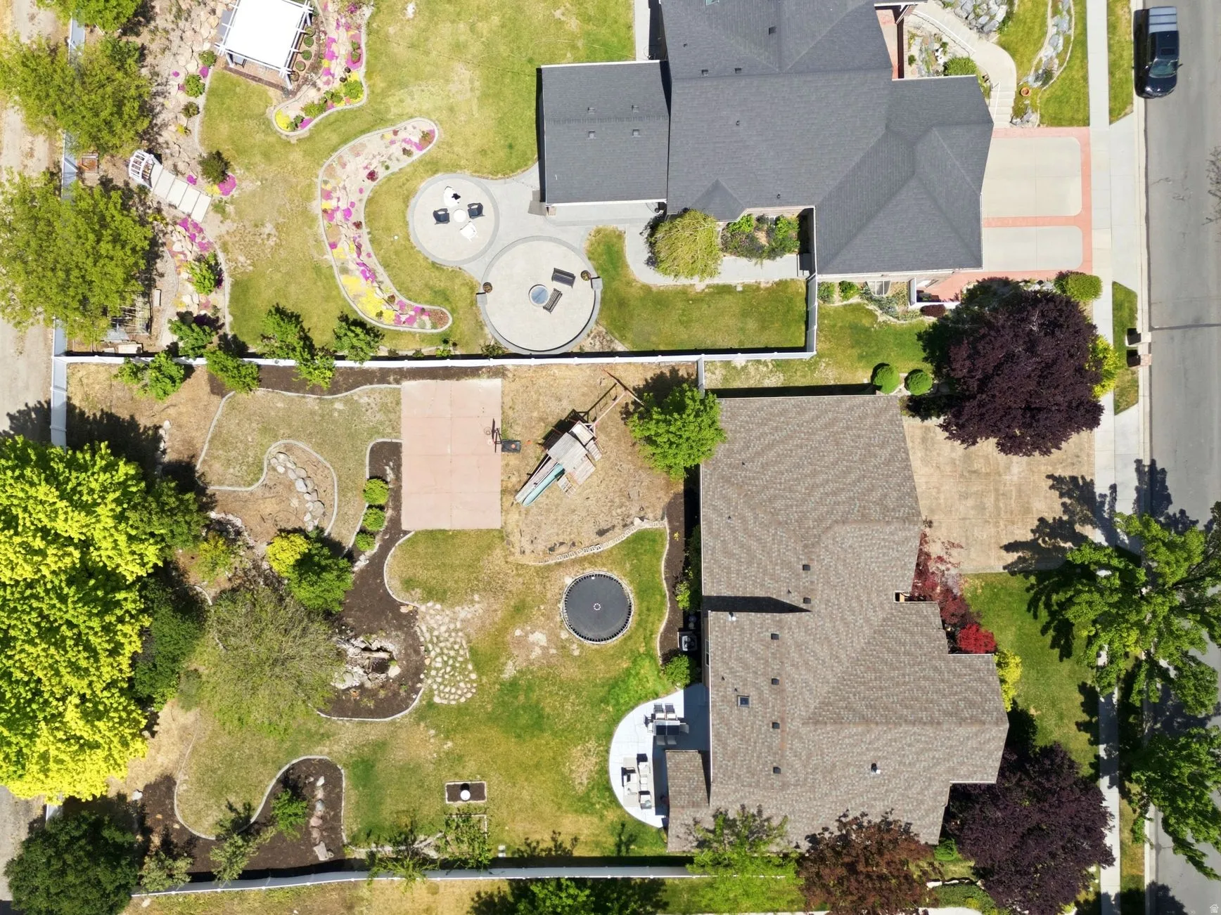 Expansive backyard featuring a manicured lawn, established trees, a dedicated sports court, a paved patio area, and garden beds with decorative stones