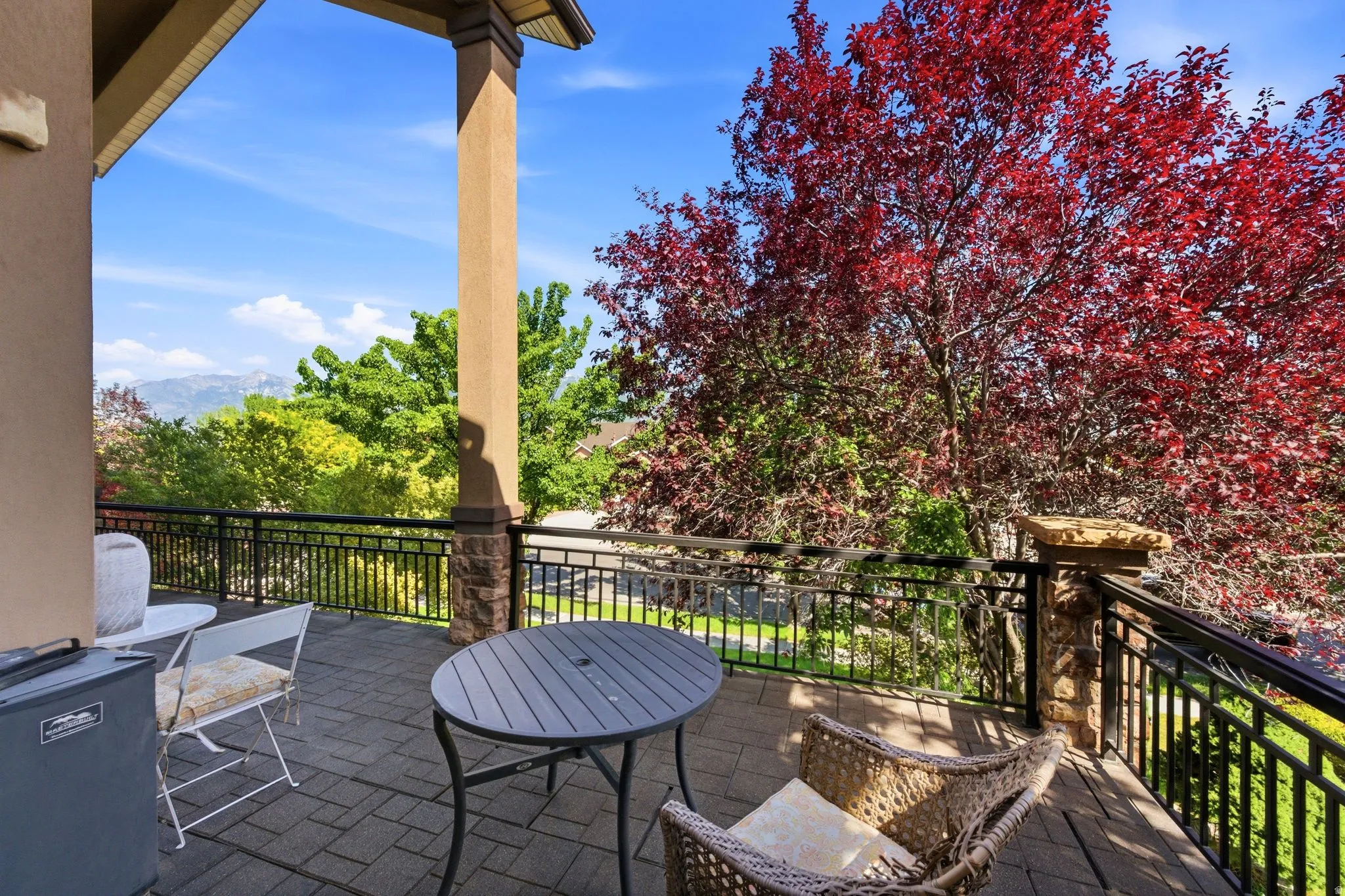 Spacious outdoor balcony with paver flooring, black wrought iron railing, and substantial stone-clad pillars, offering tree-filled scenery and distant mountain peaks