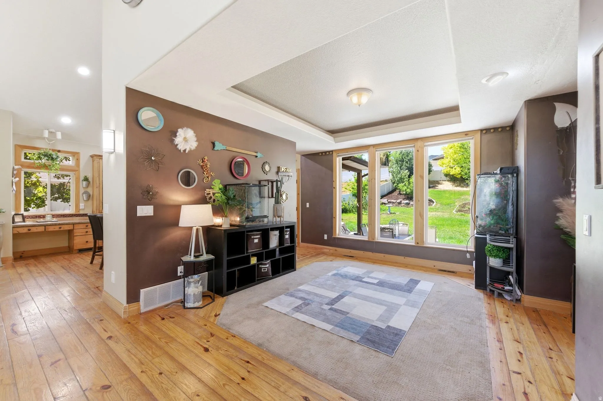 Expansive living area featuring light wood-finish flooring, a tray ceiling, and a large triple-pane window overlooking a manicured backyard