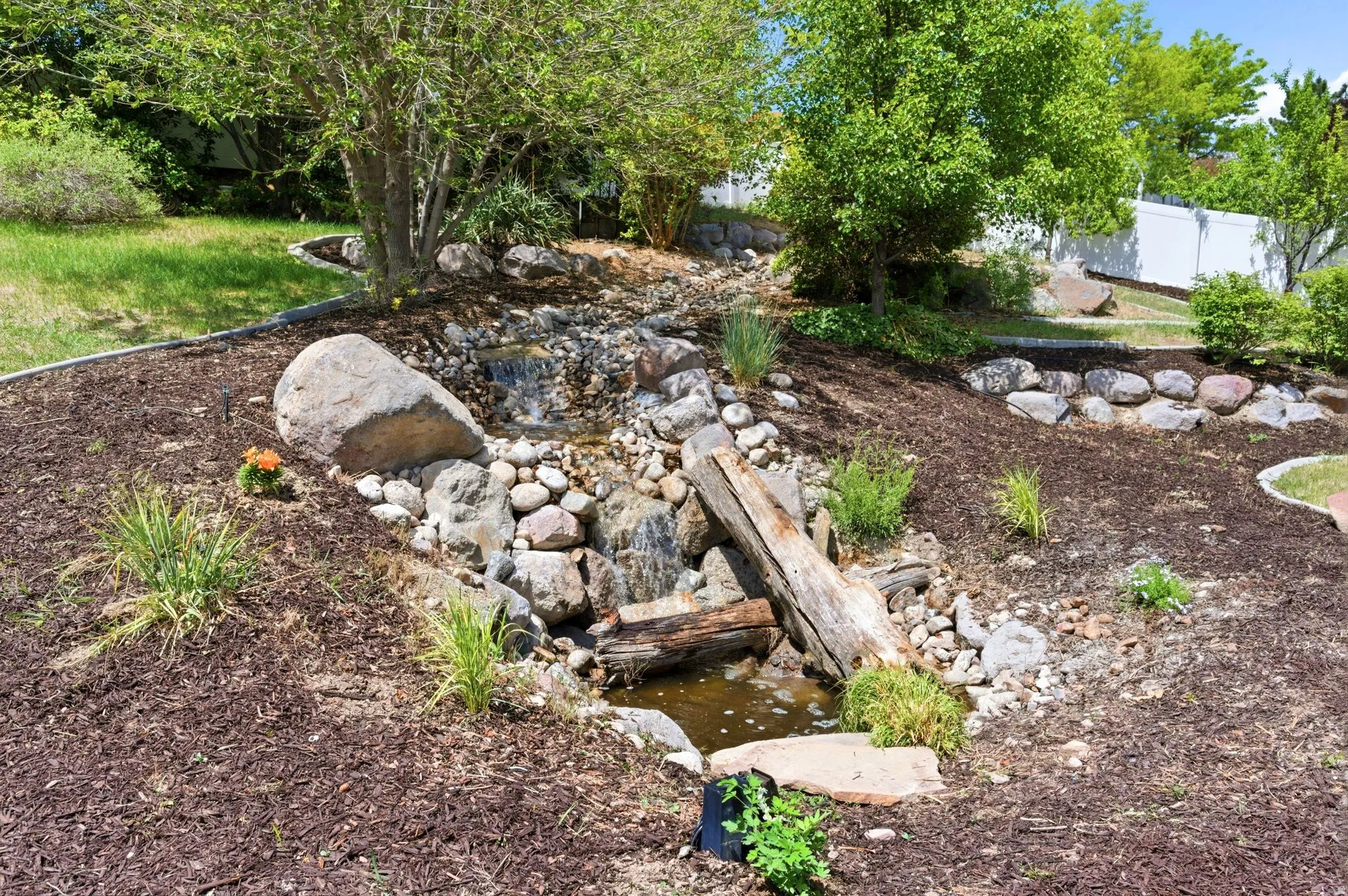 Landscaped yard featuring a multi-tiered rock waterfall, mature trees, natural boulders, bark mulch, and a white fence perimeter