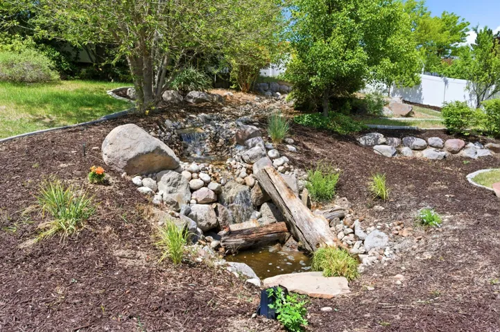 Landscaped yard featuring a multi-tiered rock waterfall, mature trees, natural boulders, bark mulch, and a white fence perimeter