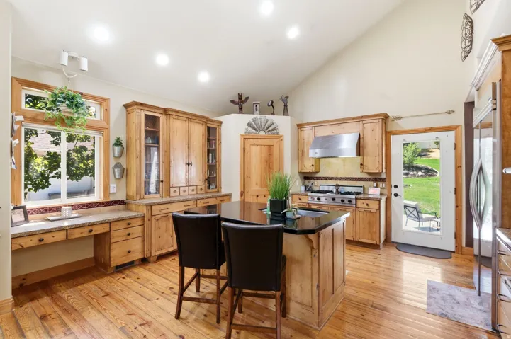 Kitchen featuring natural wood cabinetry, wood-finish flooring, a black countertop island with seating, a stainless steel range hood, and a built-in desk area