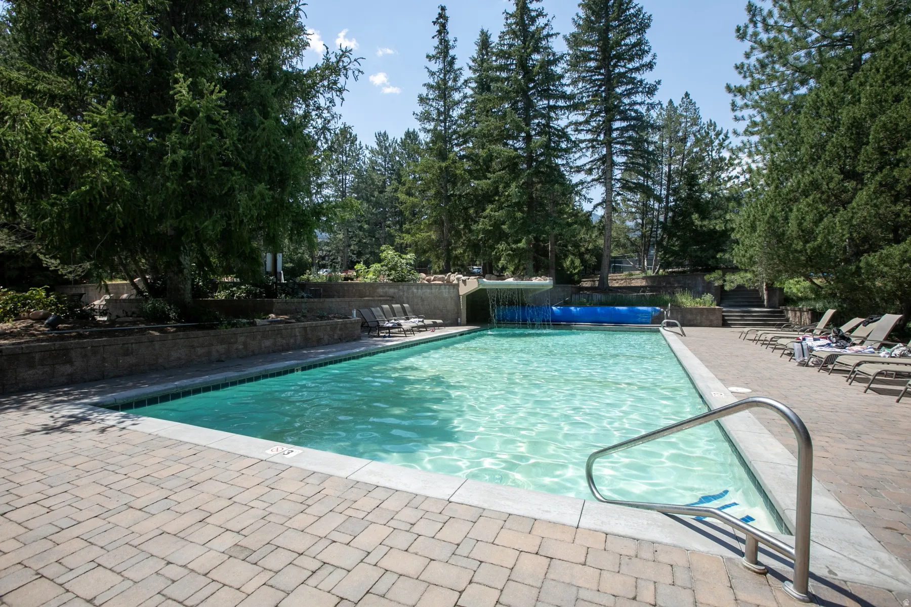 Outdoor swimming pool with integrated waterfall feature, surrounded by paver patio decking and mature trees
