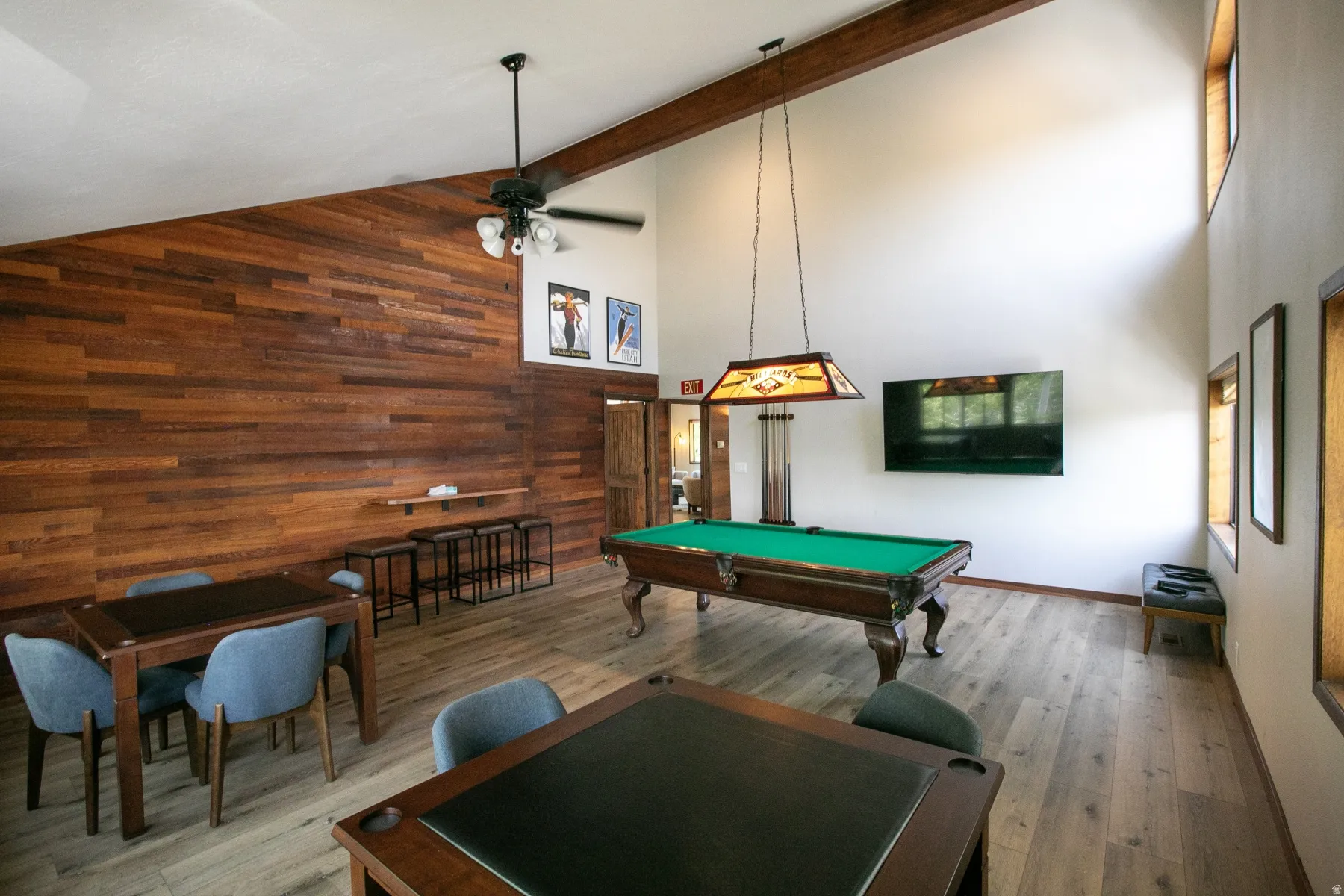 Vaulted ceiling entertainment space featuring wood-finish flooring, a full wood accent wall, exposed beam, recessed lighting, and multiple windows providing natural light
