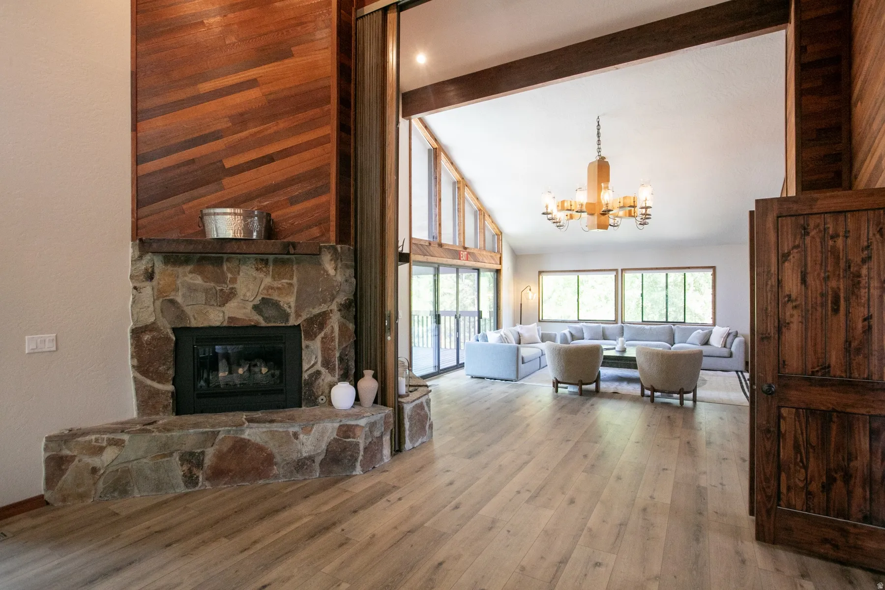 Stone-faced fireplace with a wood-paneled accent wall, light wood-finish flooring, and a vaulted ceiling with exposed beams