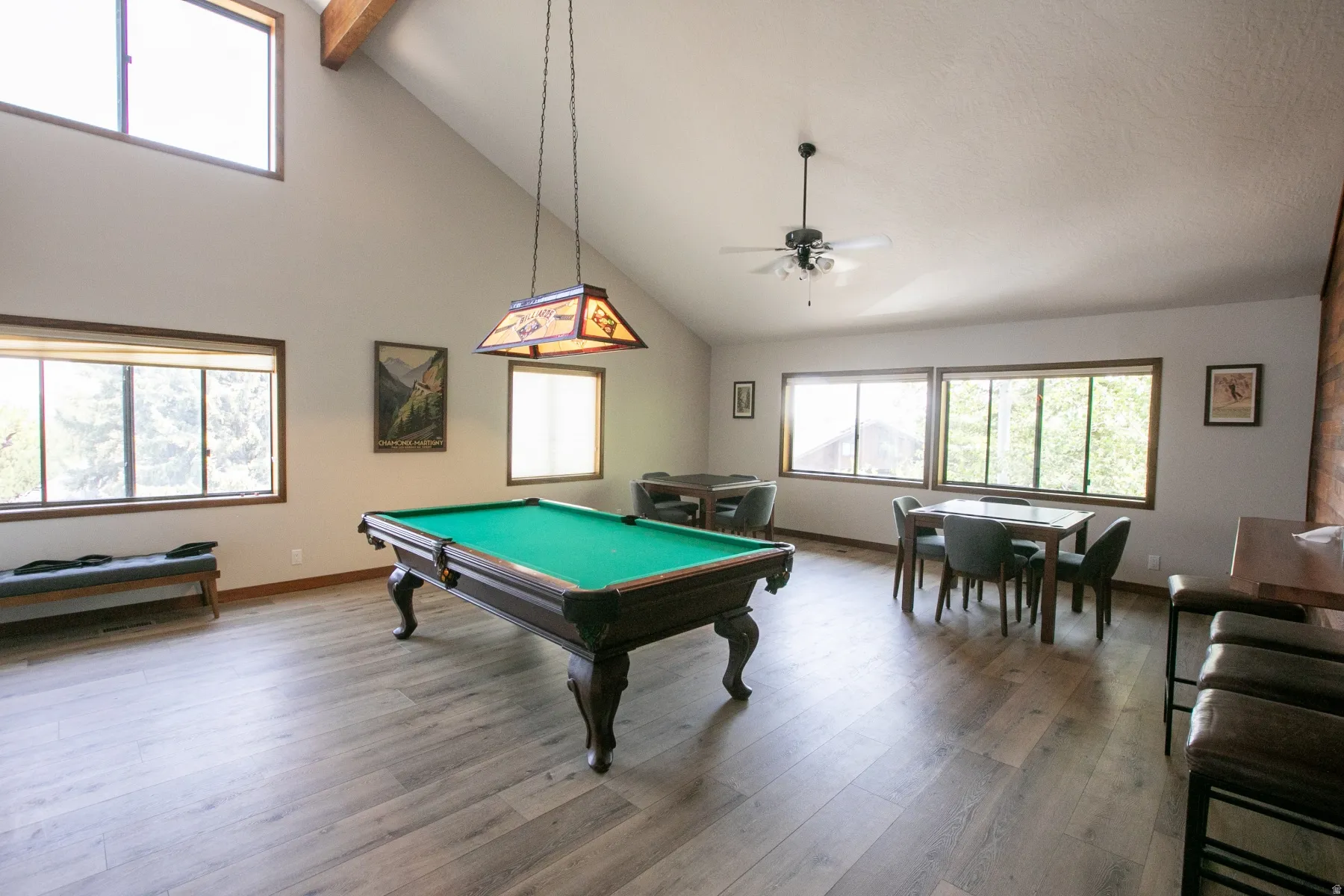 Spacious room featuring vaulted ceilings with exposed wood beams, extensive window placements, wood-finish flooring, and a ceiling fan