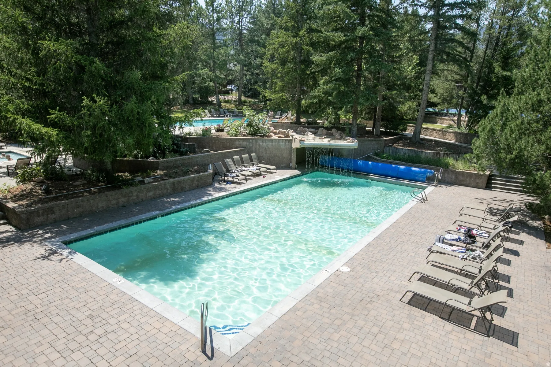 Outdoor swimming pool with brick paver surround, integrated waterfall feature, and tiered landscaping