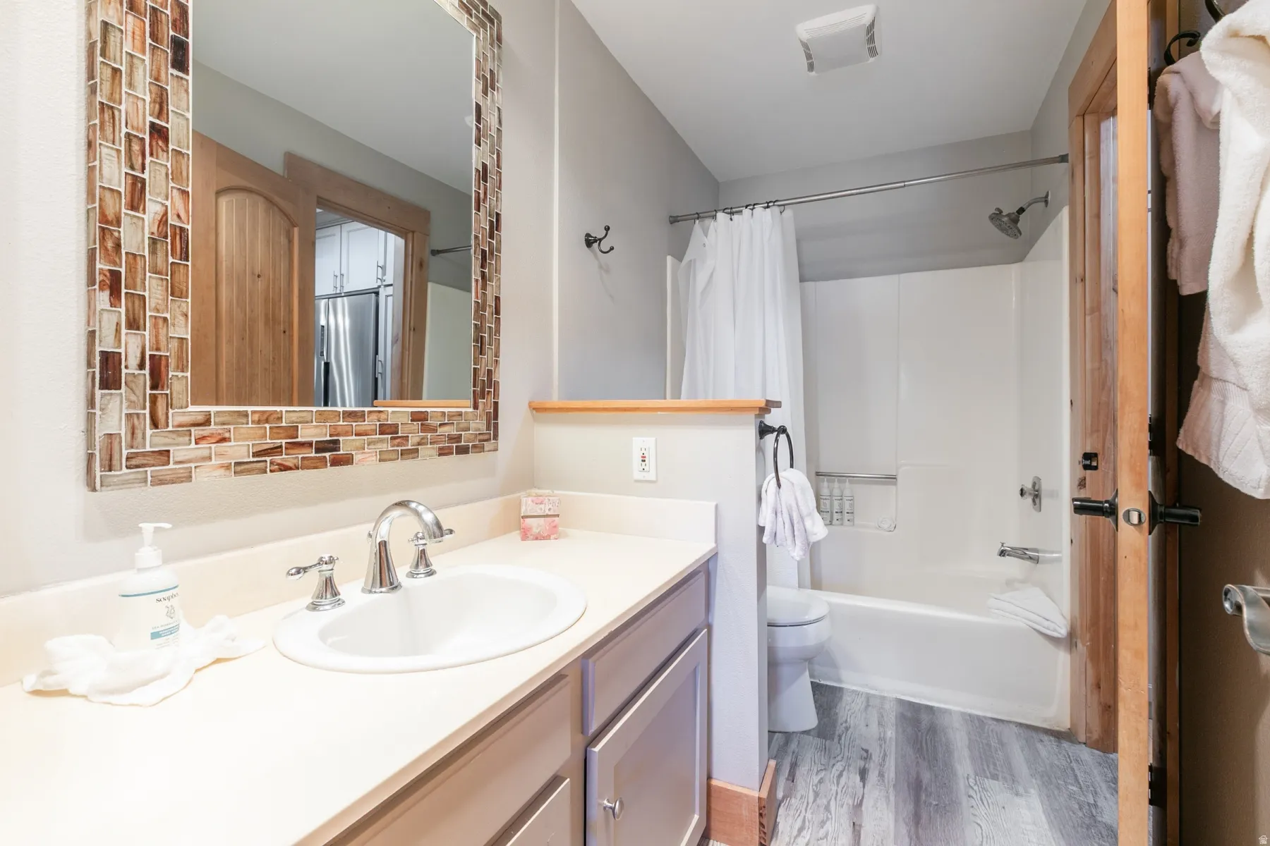 Bathroom featuring a vanity with an integrated sink and chrome fixtures, a mosaic tile-framed mirror, a full-size bathtub with shower, and wood-finish flooring