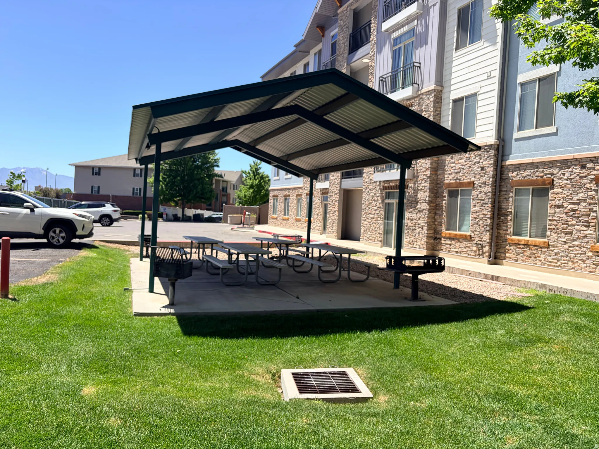 Covered outdoor amenity area featuring multiple picnic tables, a built-in barbecue grill, a concrete patio, and a manicured lawn