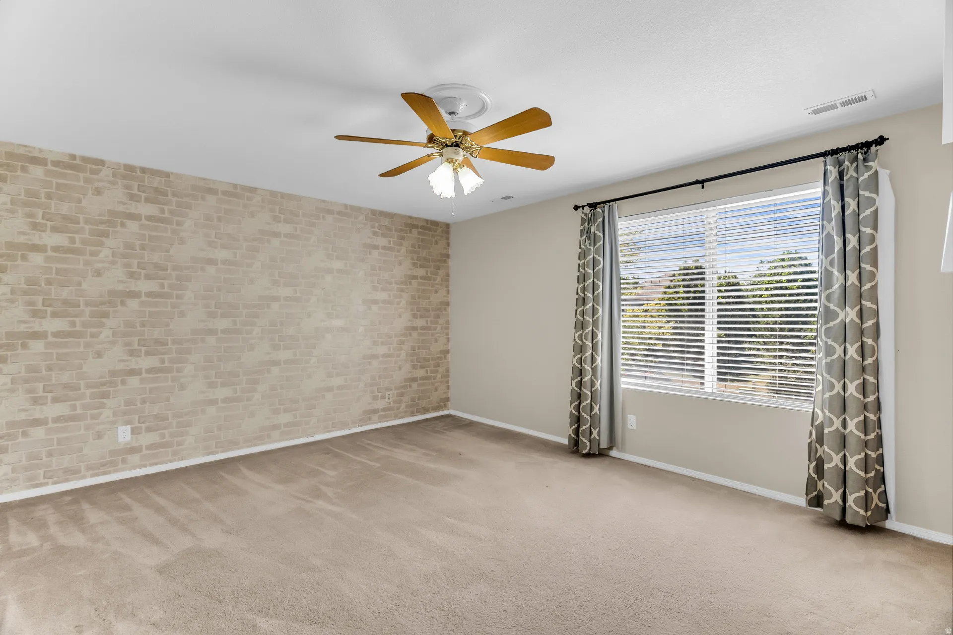 Spacious room featuring neutral carpet flooring, a brick accent wall, wood-finish ceiling fan with integrated lighting, large windows with plantation shutters and patterned drapery, and soft overhead lighting