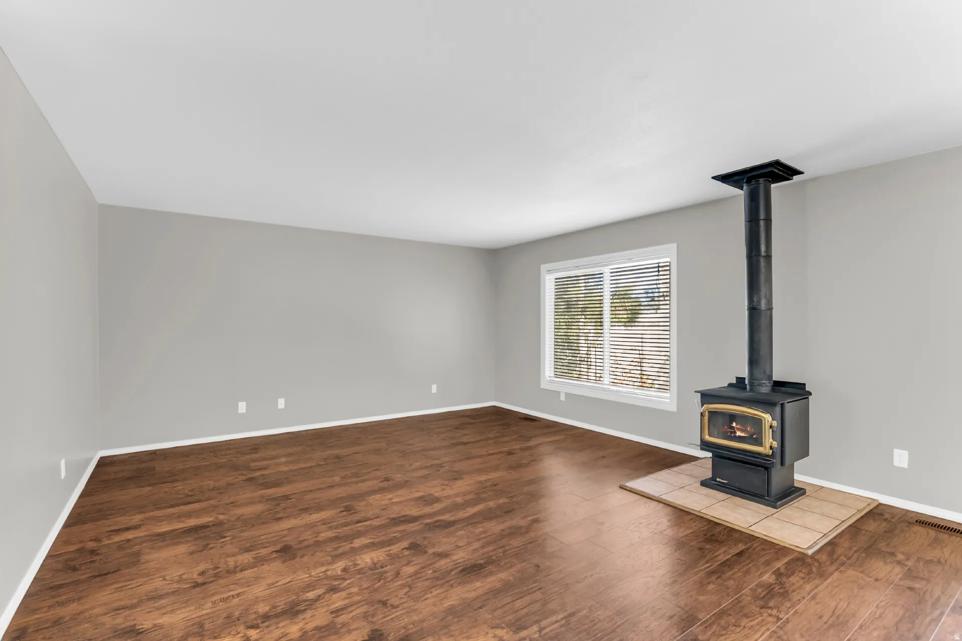 Expansive interior featuring wood-finish flooring, a modern wood-burning stove with a tiled hearth, and large window with blinds