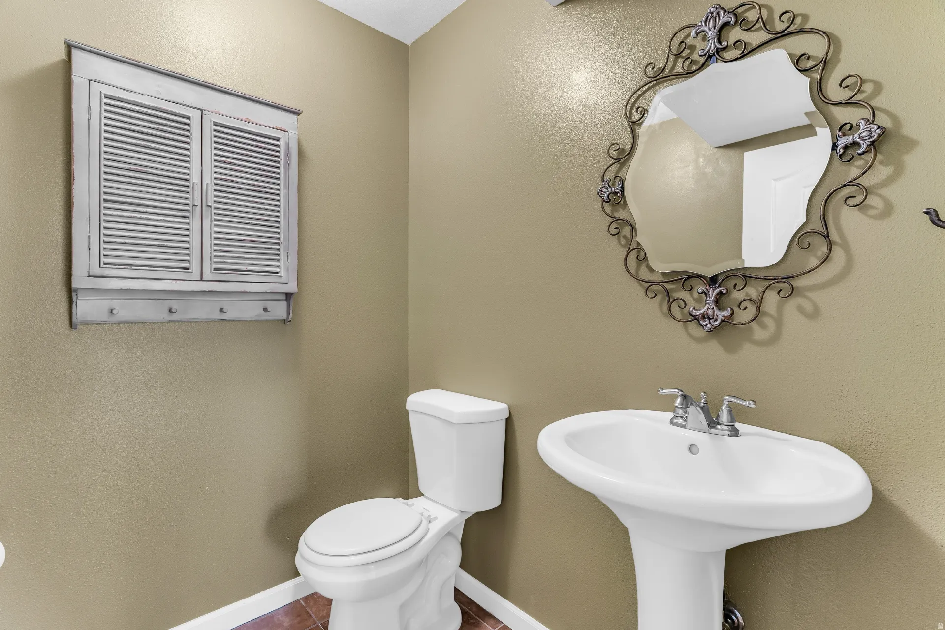 Pedestal sink with chrome faucet, toilet, wall-mounted cabinet with louvered doors and hooks, and a decorative mirror