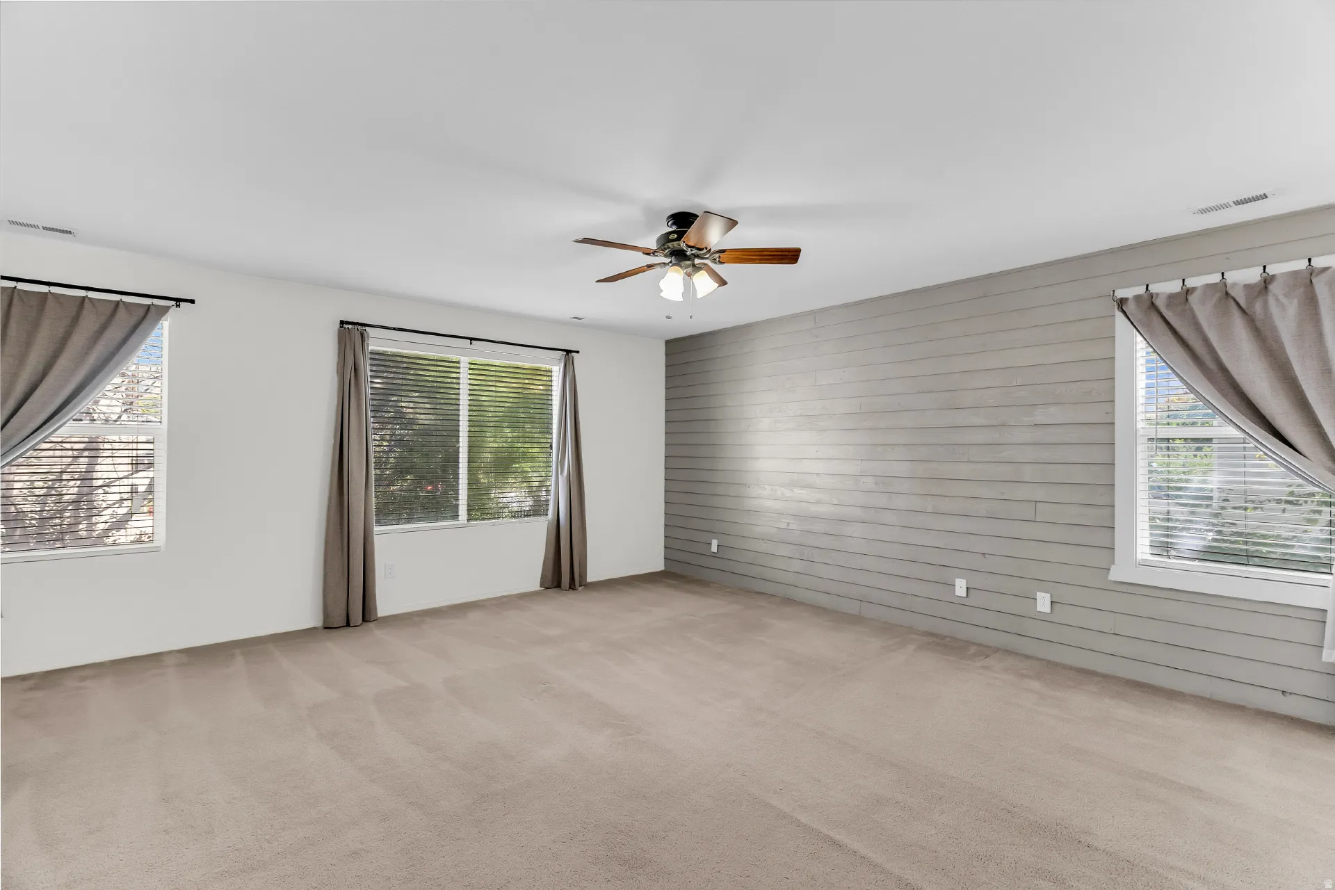 Spacious room with plush carpeting, multiple windows providing natural light, a distinctive shiplap accent wall, and a ceiling fan with integrated lighting