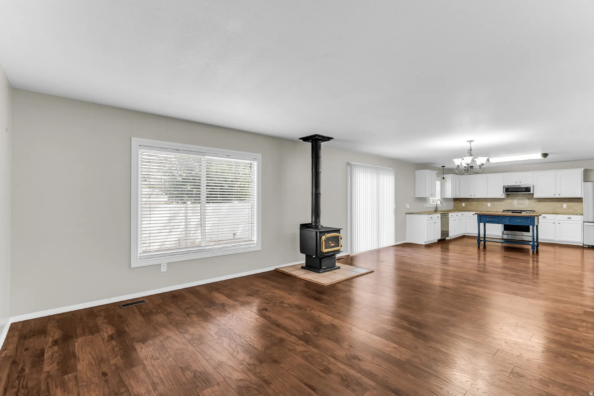 Hardwood-finish flooring, wood-burning stove with tile hearth, white window blinds, white kitchen cabinetry, and a blue kitchen island