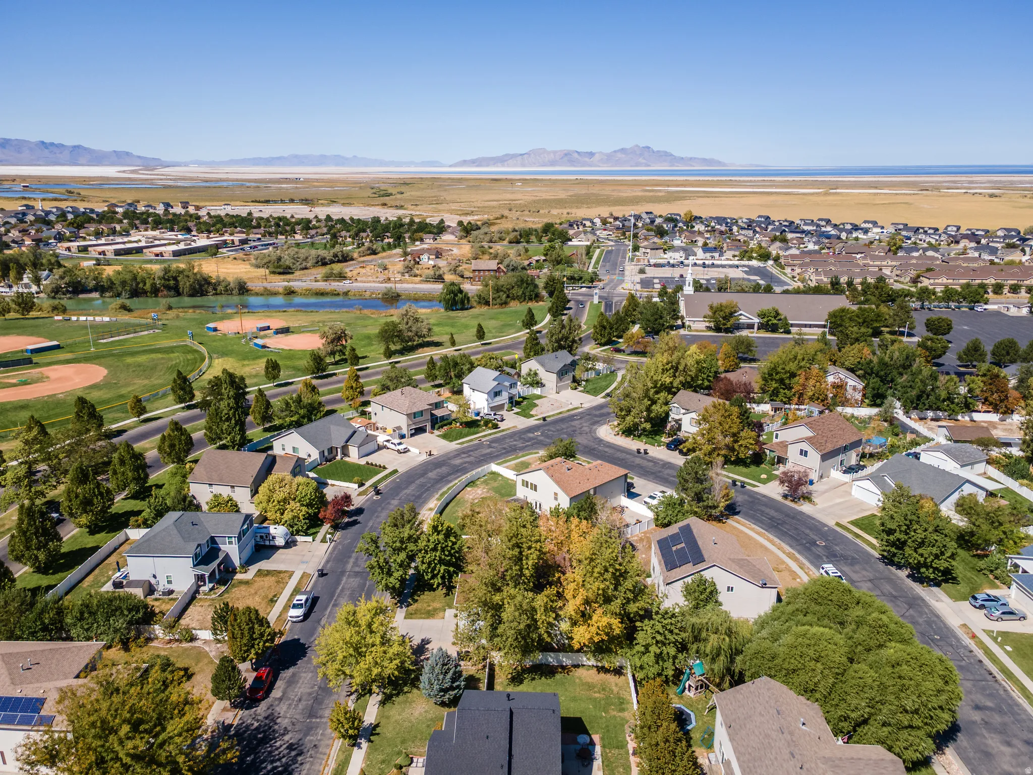 Aerial perspective showcasing a residential neighborhood with a baseball field, open water, and distant mountains