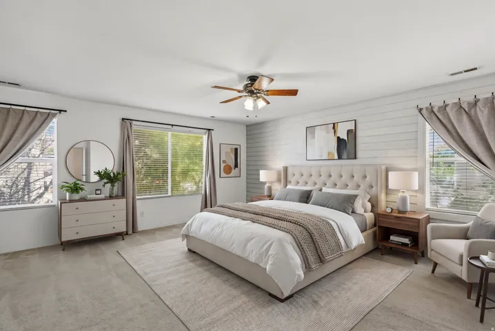 Carpeted flooring with a contrasting area rug, white textured accent wall, ceiling fan with integrated lighting, and multiple windows with blinds