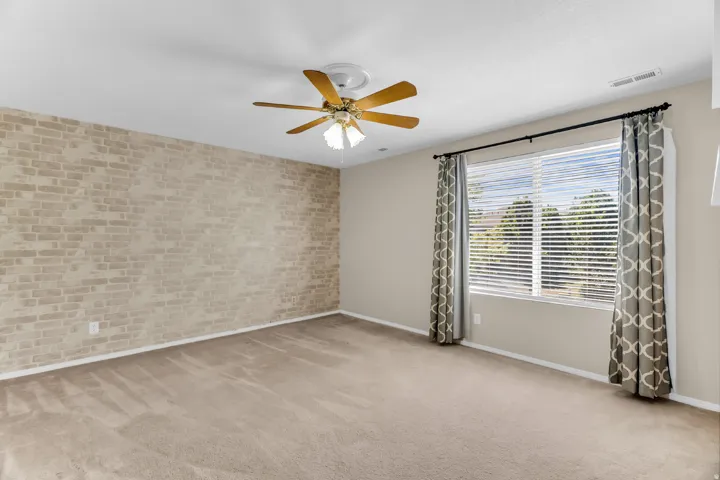 Spacious room featuring neutral carpet flooring, a brick accent wall, wood-finish ceiling fan with integrated lighting, large windows with plantation shutters and patterned drapery, and soft overhead lighting