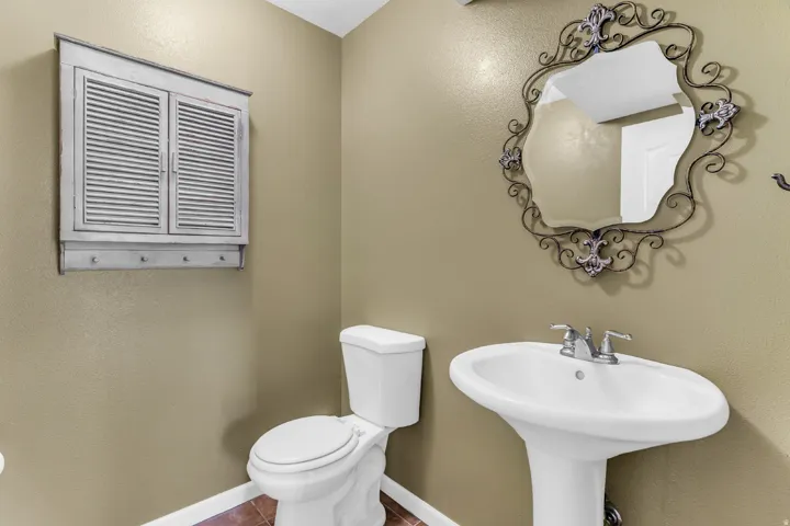 Pedestal sink with chrome faucet, toilet, wall-mounted cabinet with louvered doors and hooks, and a decorative mirror
