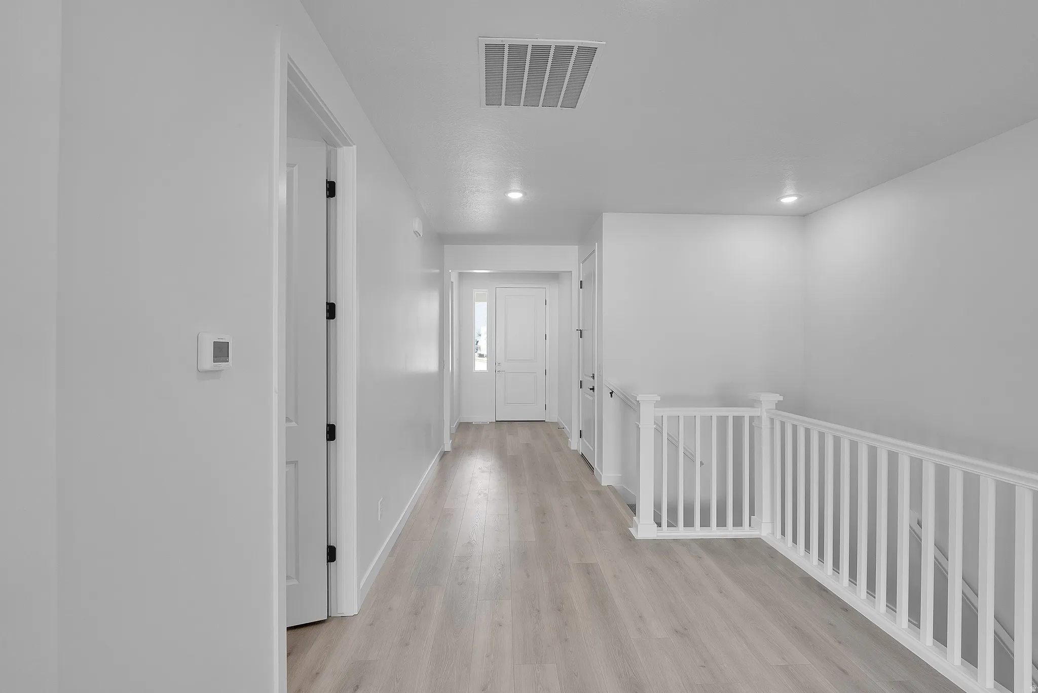 Spacious hallway with light wood-finish flooring, recessed lighting, white walls, white doors with dark hardware, and a white staircase railing