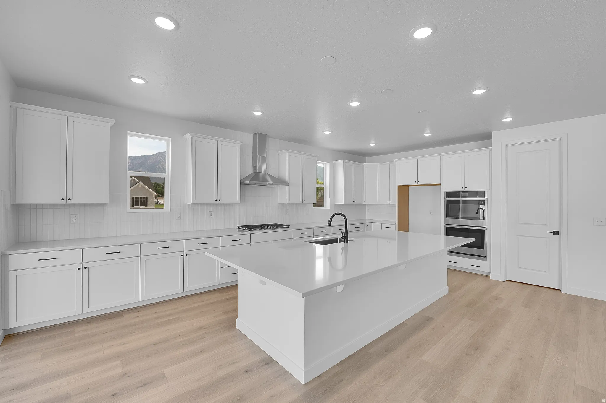 Spacious kitchen featuring an expansive island with an integrated sink, white shaker cabinetry, light wood-finish flooring, a stainless steel range hood, and double wall ovens