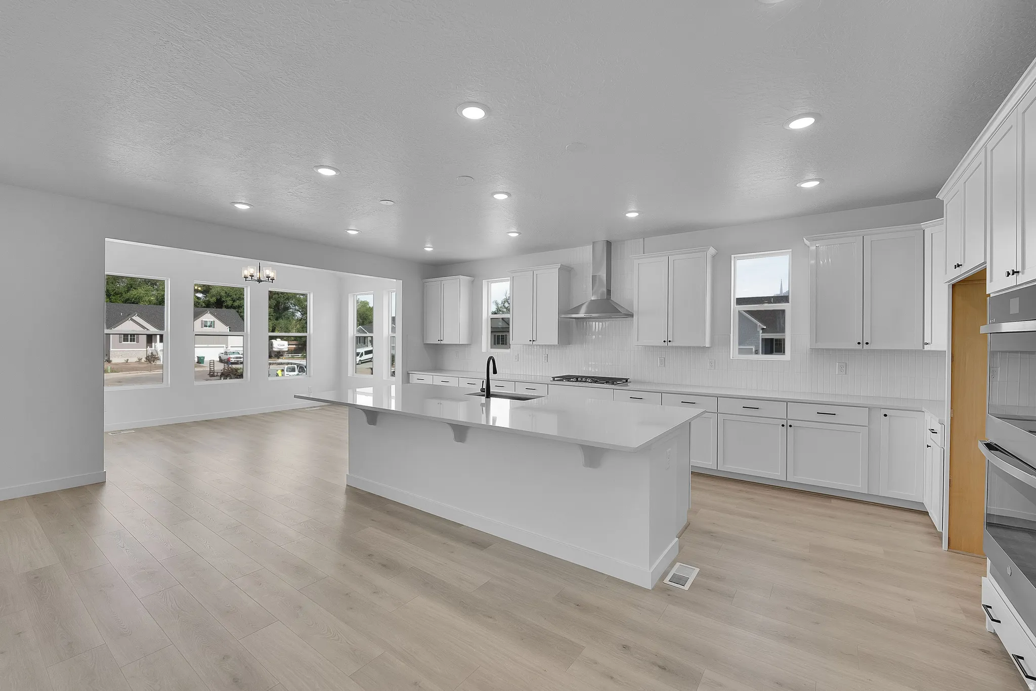 Open-concept kitchen with a large island, white shaker cabinetry, stainless steel range hood, built-in double oven, and wood-finish flooring
