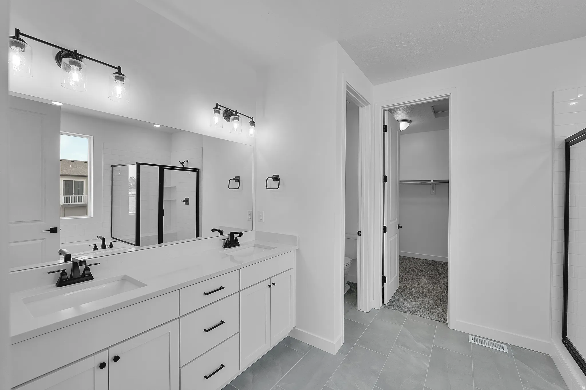 Bathroom with dual vanity featuring white cabinetry and countertops, black fixtures, a walk-in shower with black framing, and light-toned tile flooring