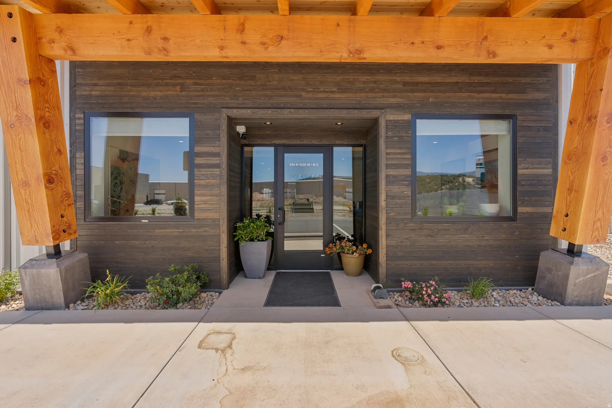 Modern building entrance featuring a glass door, two large windows, dark wood-finish siding, and exposed wood beams